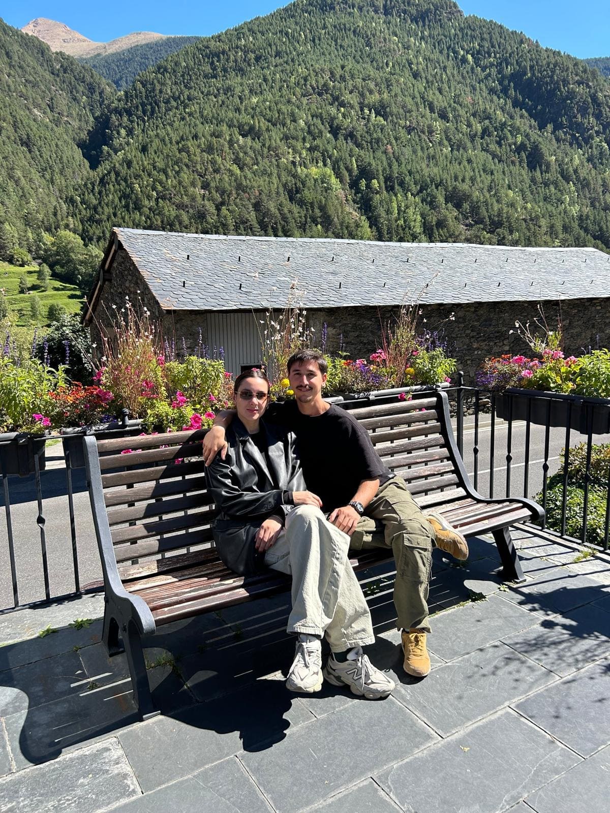 Pareja sentada en un banco rodeada de flores y montañas, inspirando paisajismo y jardinería, ideal para amantes de la naturaleza y la venta de plantas